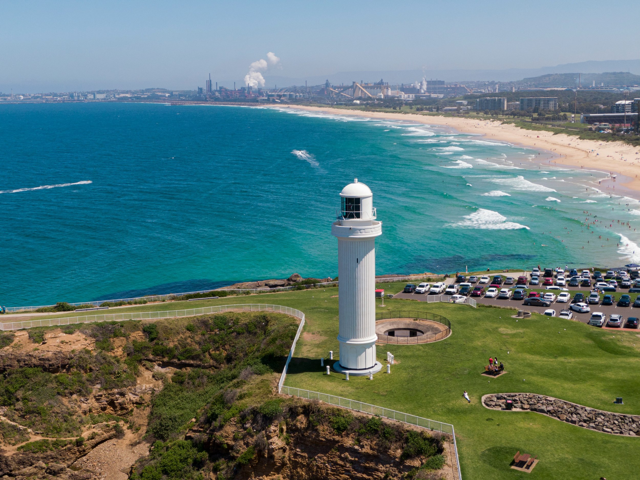 A lighthouse on a grassy area near a beach with people, a parking lot with cars, and in the background an industrial area with factories emitting smoke.