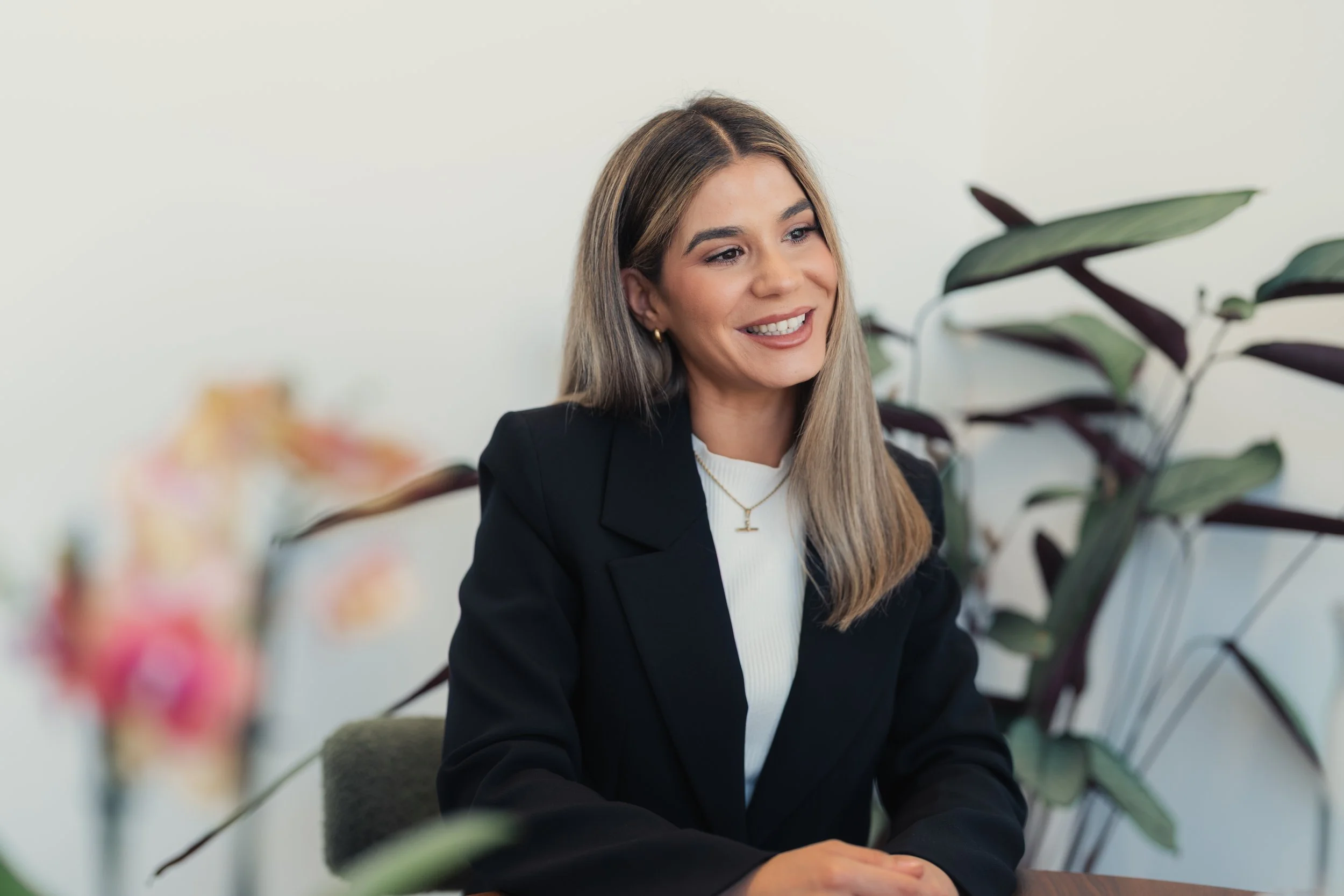 A young woman with long blonde hair, wearing a black blazer over a white top, sitting at a table with blurred pink flowers in the foreground and green plants in the background, smiling.
