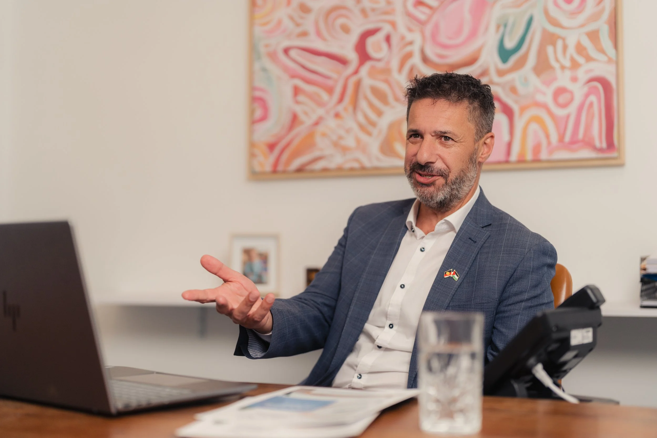 A man in a business suit gestures with his hands while speaking during a meeting in an office. There is a colorful abstract painting on the wall behind him, a laptop in front, and a water glass on the table.
