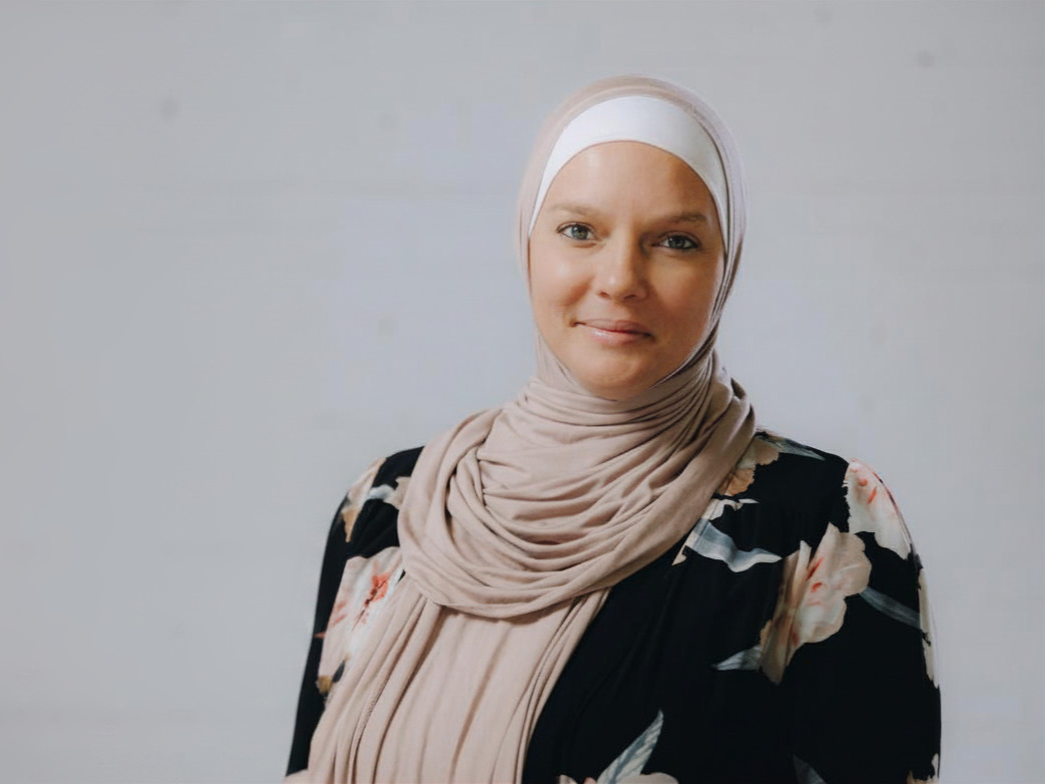 A woman wearing a beige hijab and a floral blouse standing against a plain background.