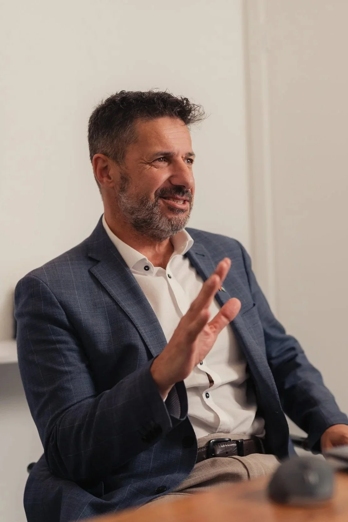A middle-aged man in a suit and white shirt is sitting and speaking, gesturing with his right hand, in a professional setting.