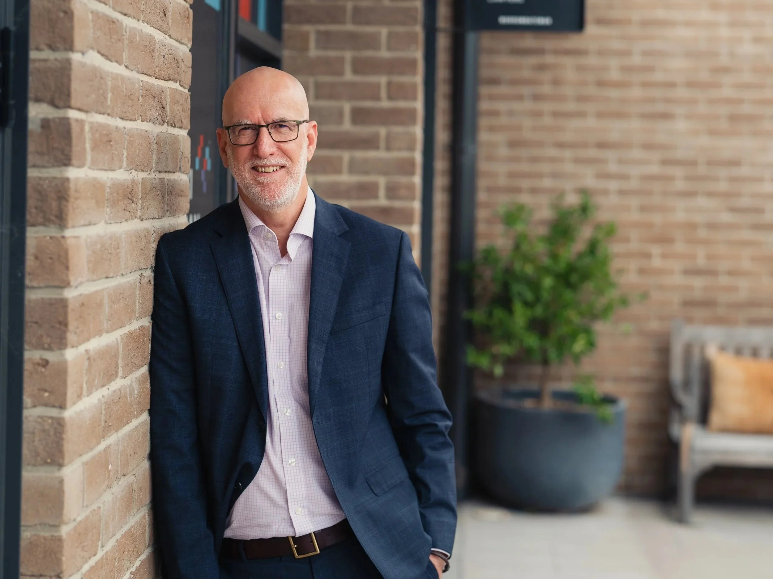 A middle-aged man with glasses, a beard, and a bald head, dressed in a dark blue blazer and pink shirt, leaning against a brick wall outside a building with a potted plant in the background.