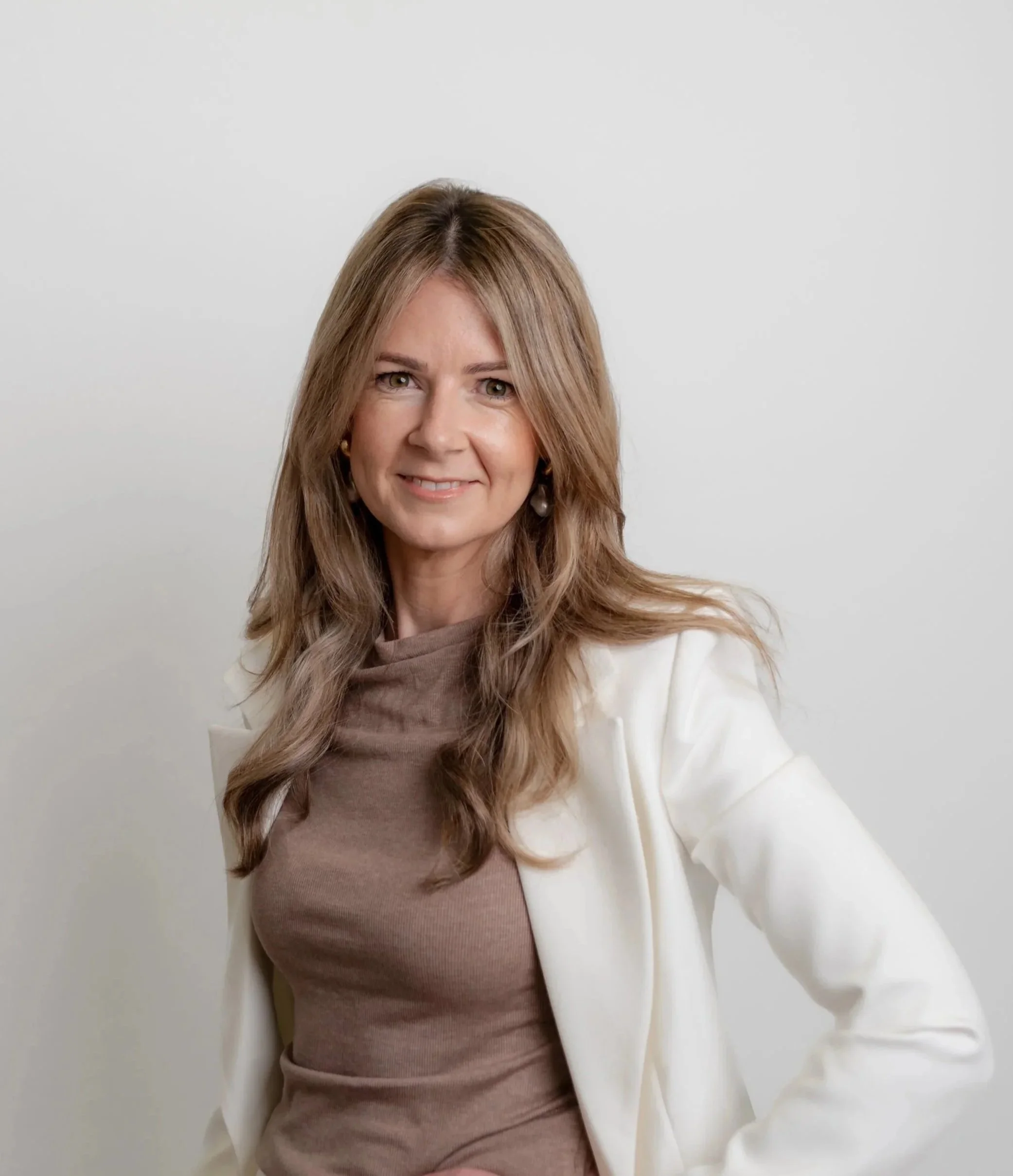A woman smiling, with light brown hair, wearing a beige top and a cream-colored blazer, standing against a plain white wall background.