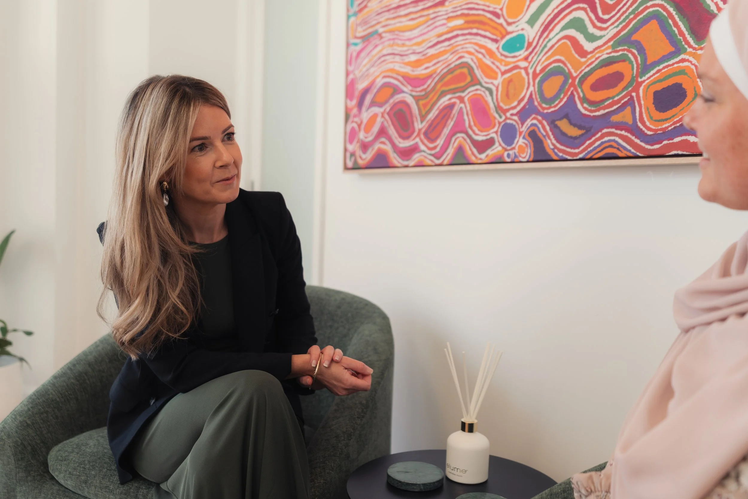 Two women engaged in conversation in a modern setting, with colorful abstract art on the wall and a white bottle with sticks on a table between them.