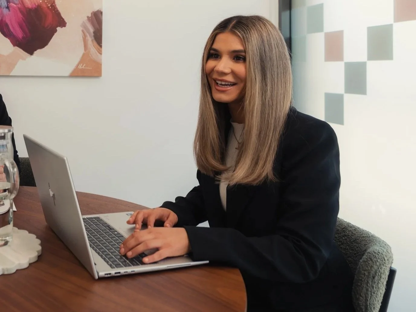A woman with long blonde hair working on a laptop at a conference table, smiling, in a modern office setting.