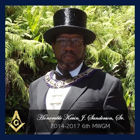 Man dressed in formal suit with a top hat, glasses, and a Masonic regalia, standing outdoors with lush greenery in the background.