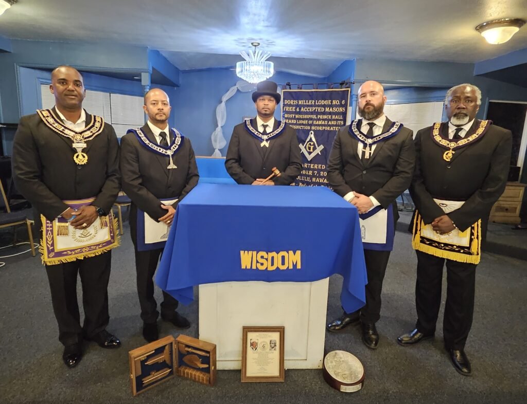 Five men in formal suits and Masonic regalia standing behind a table draped with a blue cloth that has 'WISDOM' printed on it, in a room with blue walls, a chandelier, and framed certificates or documents on the wall and floor.