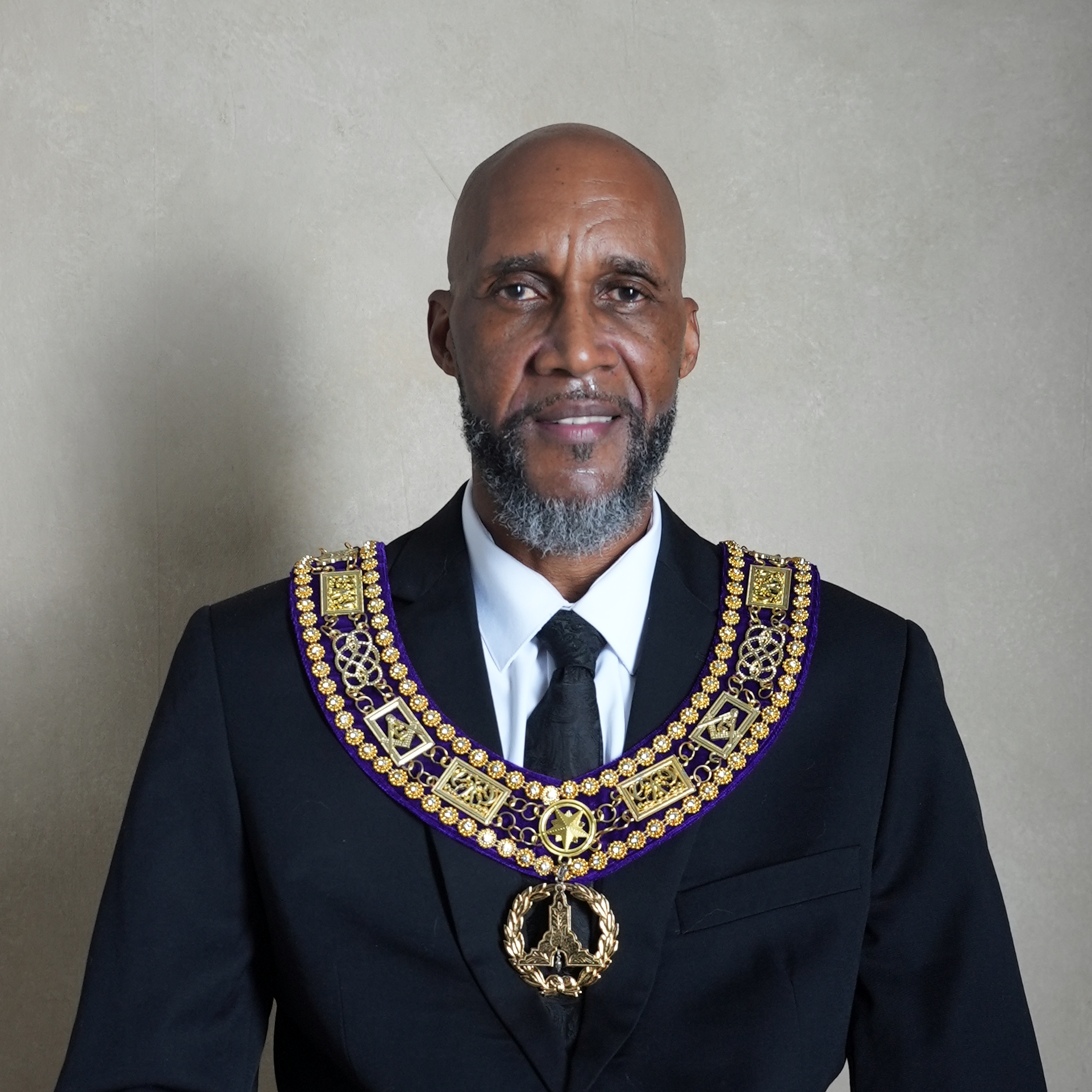 A man in a black suit with a white shirt and black tie wearing extensive gold and purple Masonic regalia, including a large collar with various symbols and a pendant, standing against a plain white background.