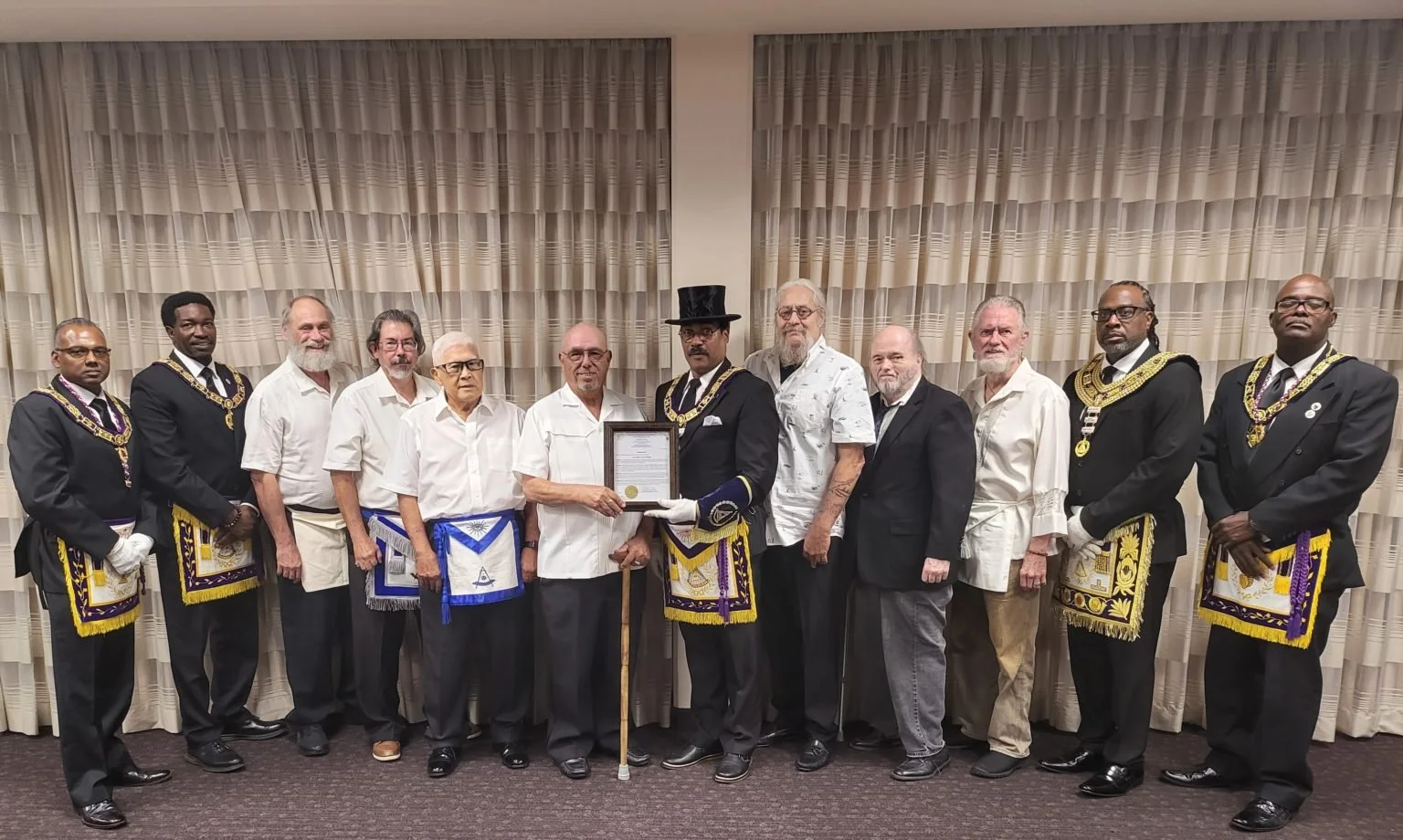 Group of men in formal and Masonic regalia standing in front of curtains, one holding a framed document.
