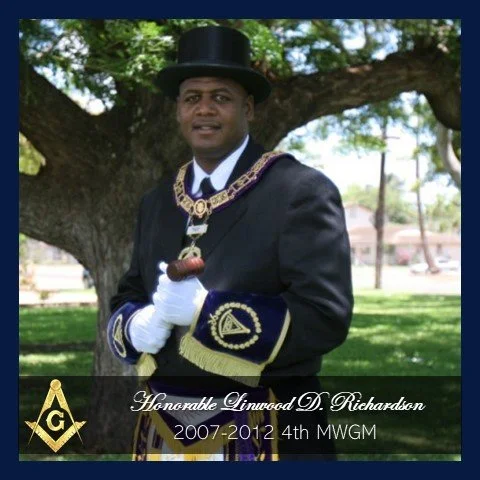 A man in Masonic regalia, wearing a black hat and suit, standing outdoors under a tree, with a Masonic apron and Jewel, holding gloves, and smiling.