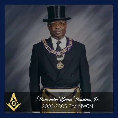 A man in formal attire with a top hat, glasses, and a ceremonial collar, standing against a gray background.