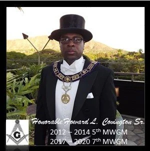 Man wearing a black suit, top hat, and ceremonial chains standing outdoors with greenery and mountains in the background.