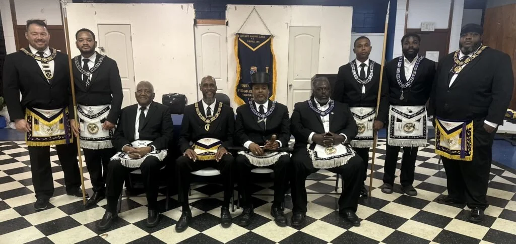 Nine men dressed in formal Masonic regalia, with aprons and sashes, posed in a Masonic lodge with checkered black and white flooring. Seven men are standing, and two are seated in the middle.