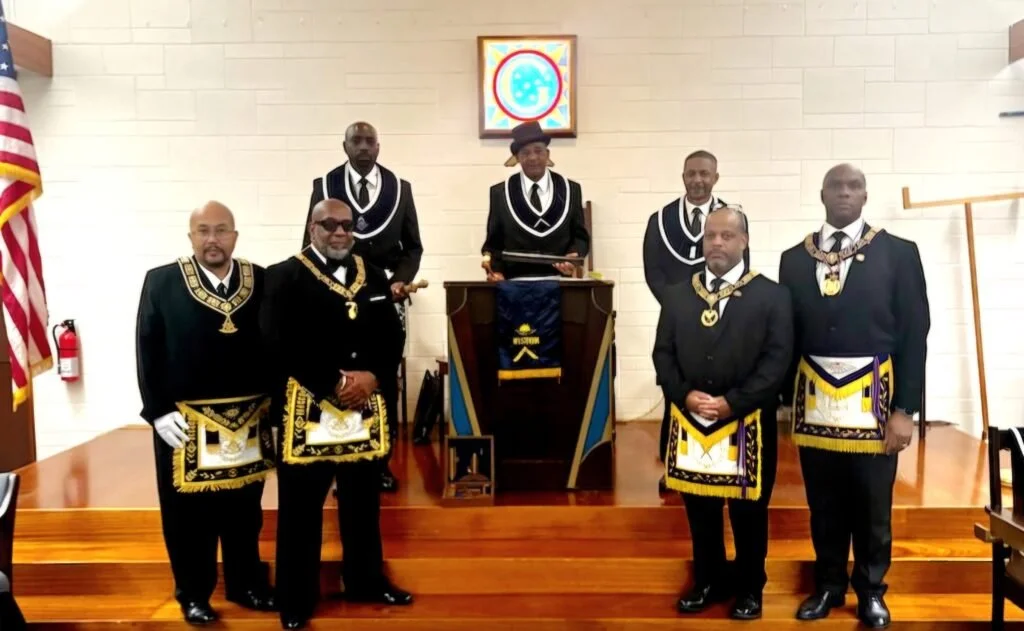 Group of seven men in formal Masonic regalia standing in a Masonic lodge room, with some wearing black suits with embroidered aprons and others with aprons and collar jewels, in front of a raised platform with a wooden podium, an American flag to the side, and a framed Masonic symbol on the wall.