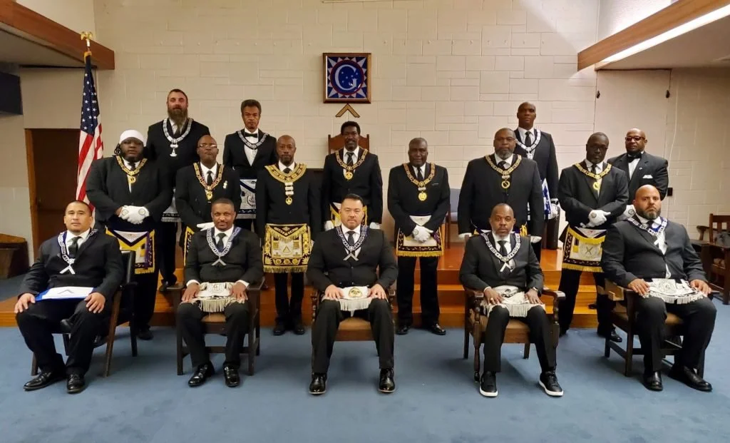 Group of men dressed in formal Masonic regalia, posing for a photo inside a Masonic lodge with a flag and Masonic symbols on the wall.