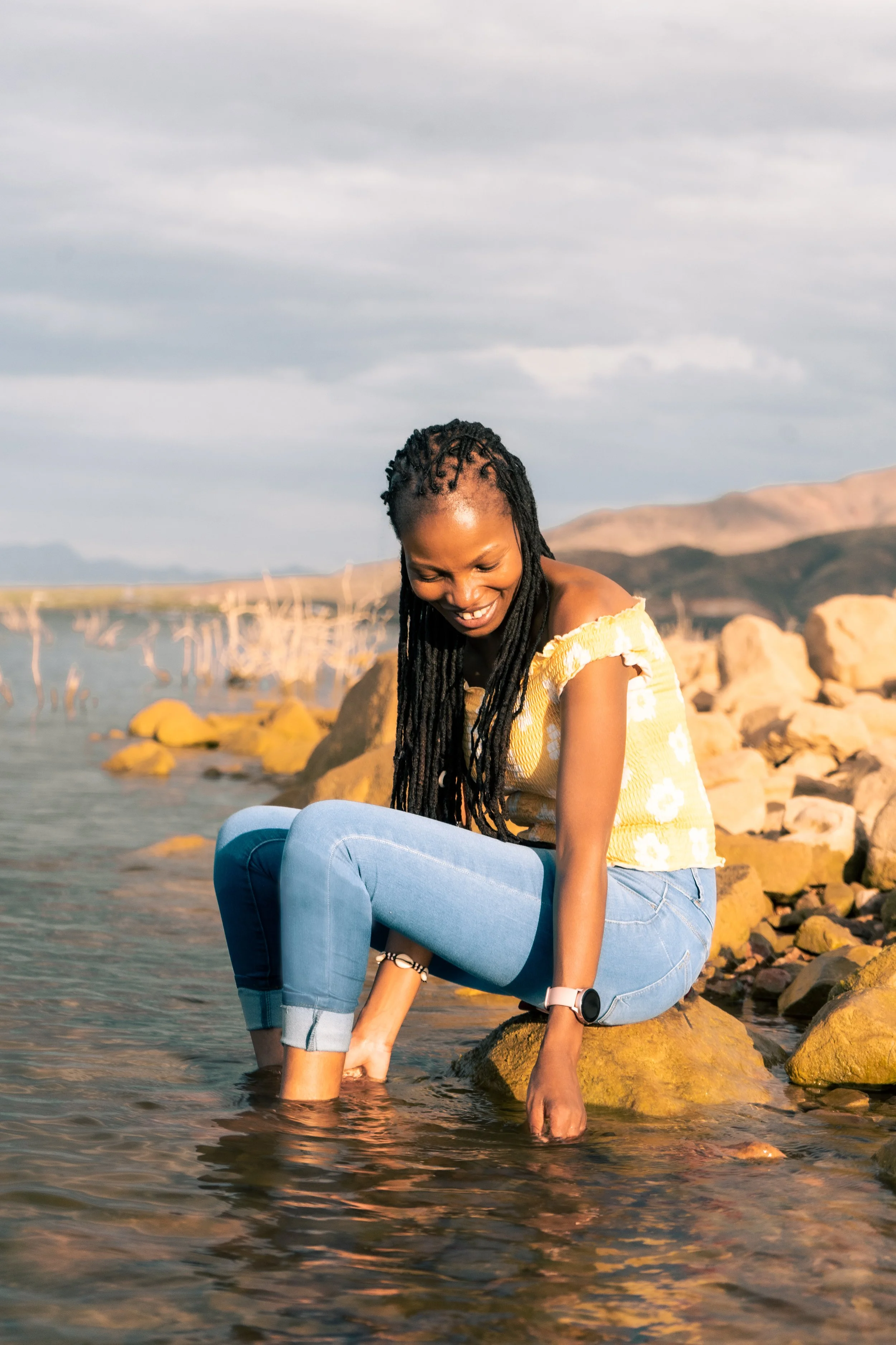A woman with long braided hair, wearing a yellow off-shoulder top with white floral patterns and blue jeans, sits by the rocks in shallow water at the beach, smiling and looking down. The background features rocks, water, mountains, and a cloudy sky.