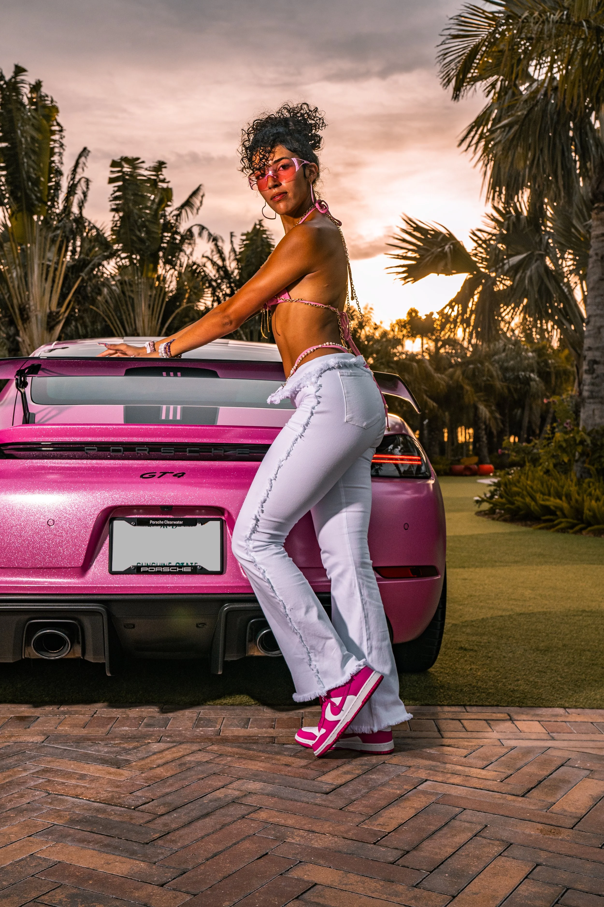 A woman with curly hair wearing pink-tinted sunglasses, a pink bikini top, and white pants with pink sneakers, stands beside a pink Porsche GT4 car in an outdoor setting with palm trees and a sunset sky in the background.