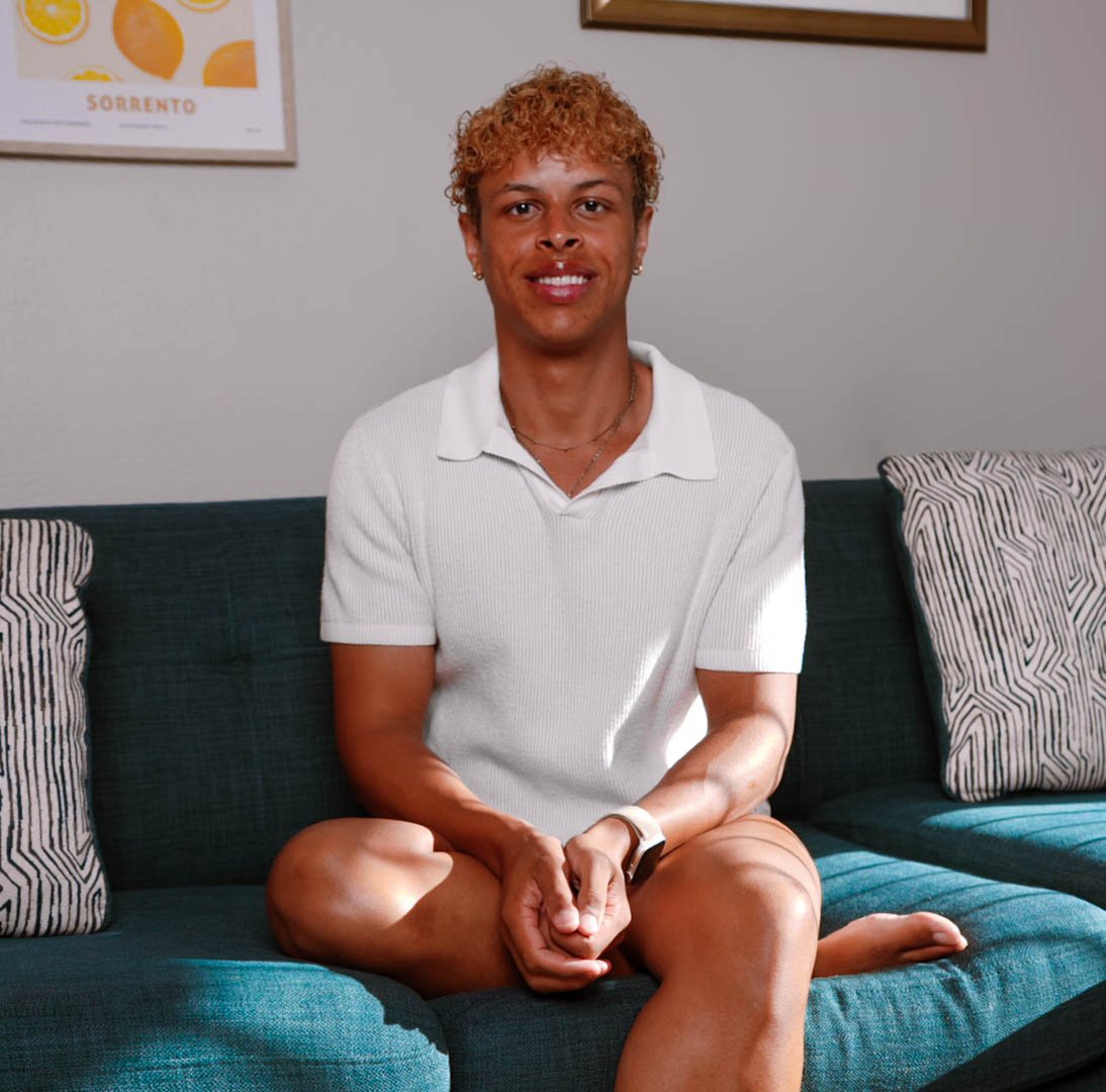 A person with short curly hair sitting cross-legged on a blue couch, smiling, wearing a white polo shirt and wristwatch, with patterned pillows and framed artwork on the wall behind them.