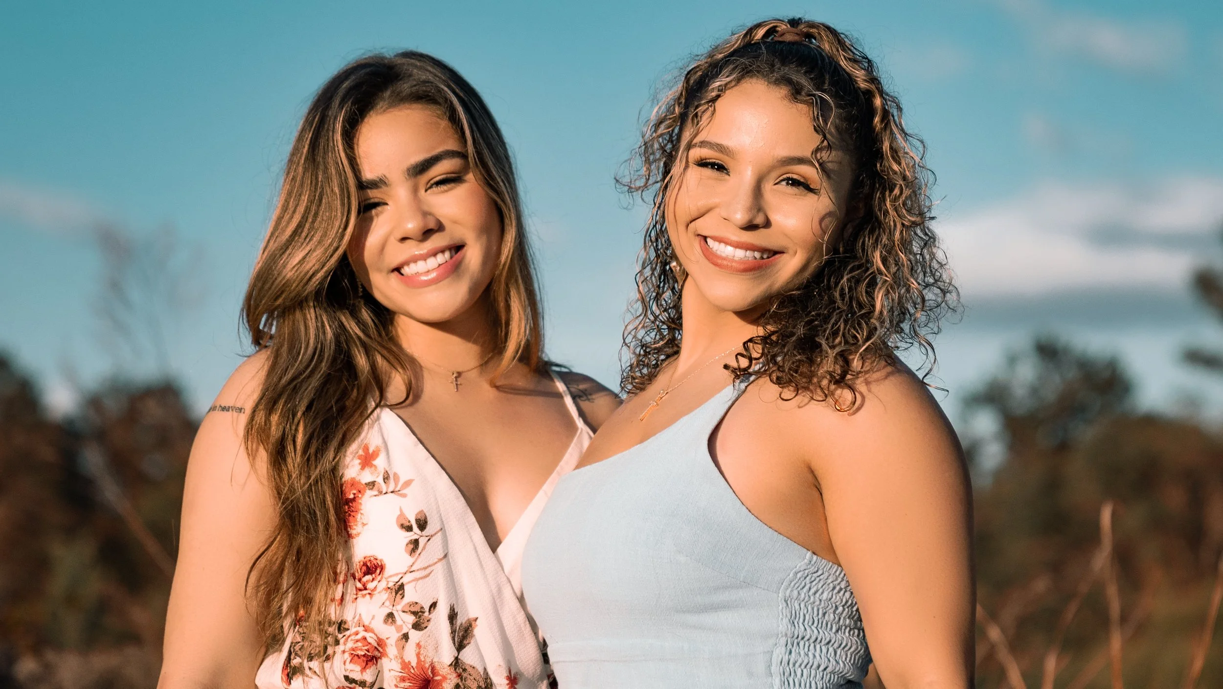 Two young women smiling outdoors on a sunny day, standing close together with a blurred landscape in the background.