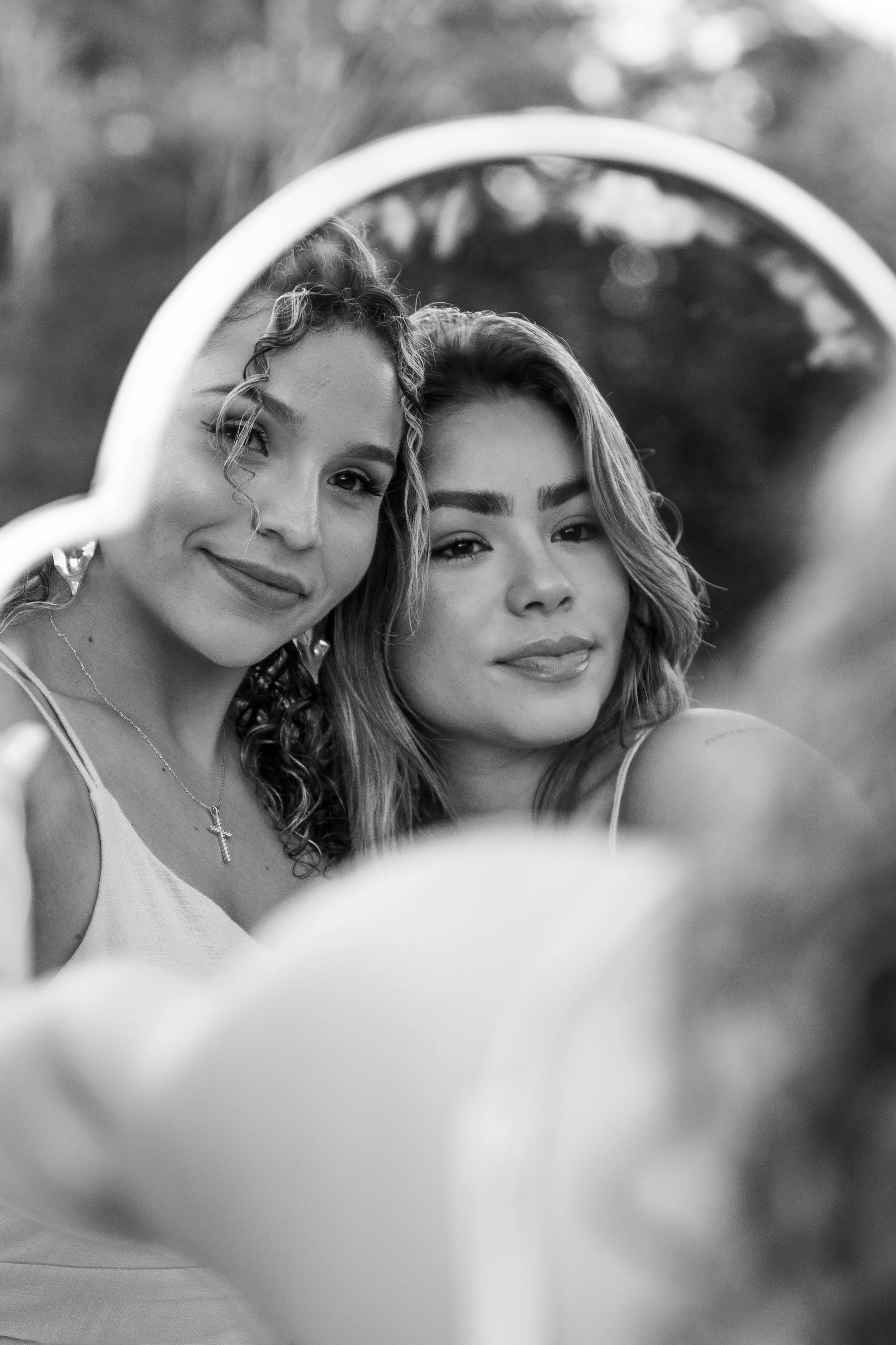 Black and white photograph of two young women taking a selfie, seen through a round mirror. Both are smiling and looking into the camera, with natural outdoor background.