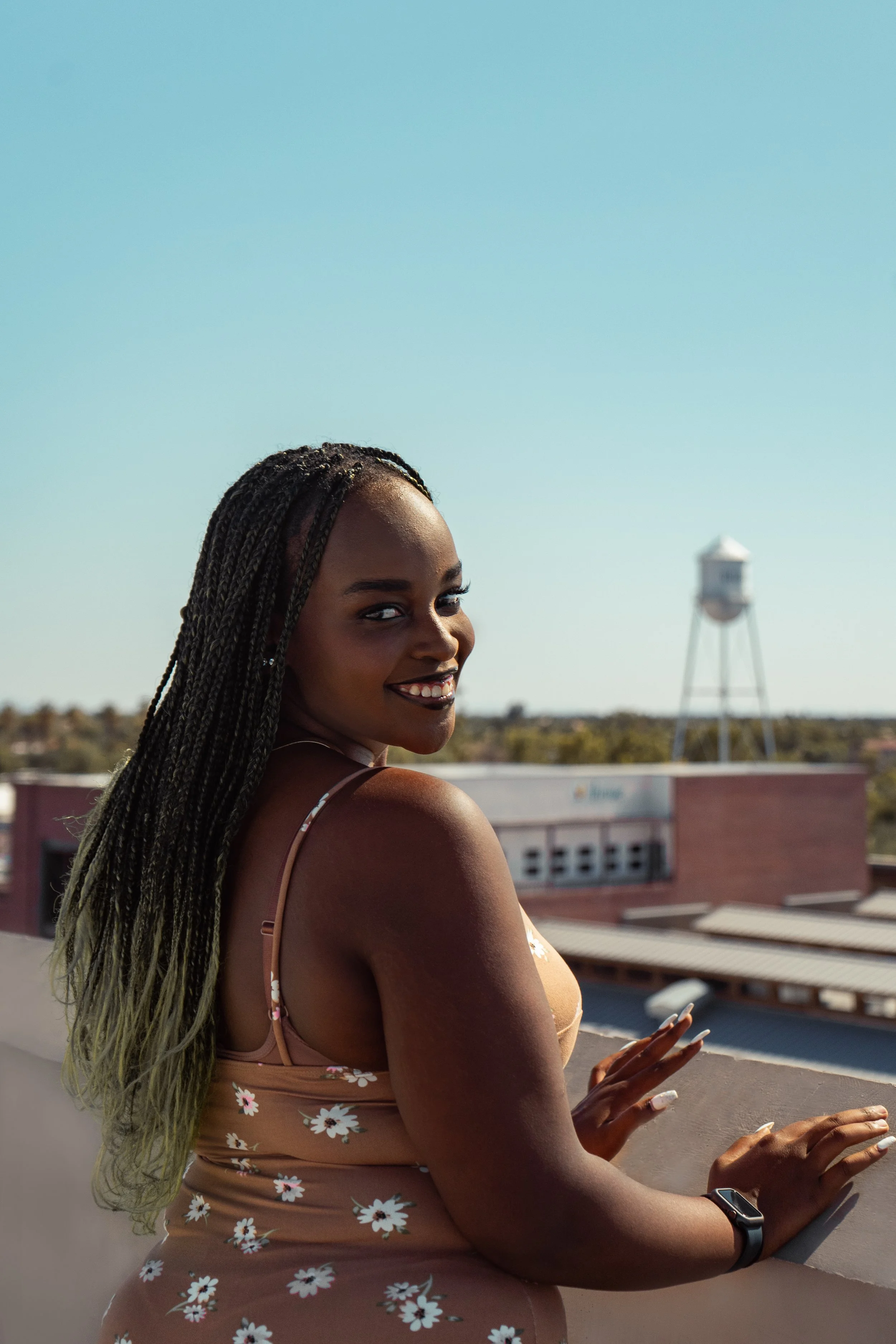 A woman with long braided hair smiling and looking over her shoulder, standing on a rooftop with a city skyline and water tower in the background under a clear blue sky.