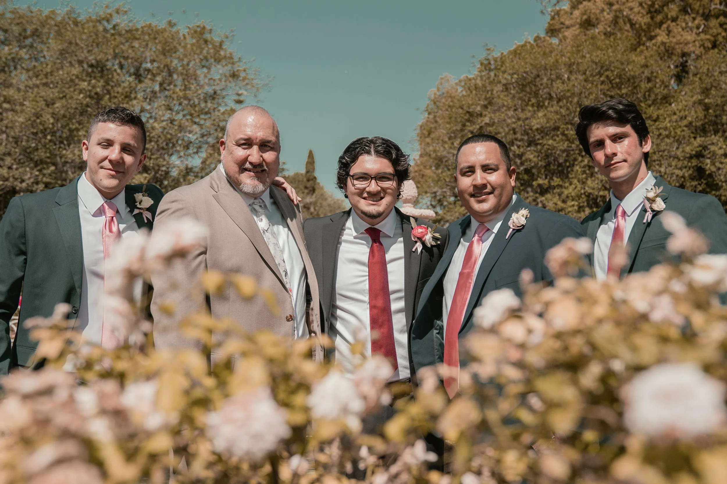 Group of five men in suits and ties standing outdoors, smiling, with trees and blue sky in the background, during daytime.