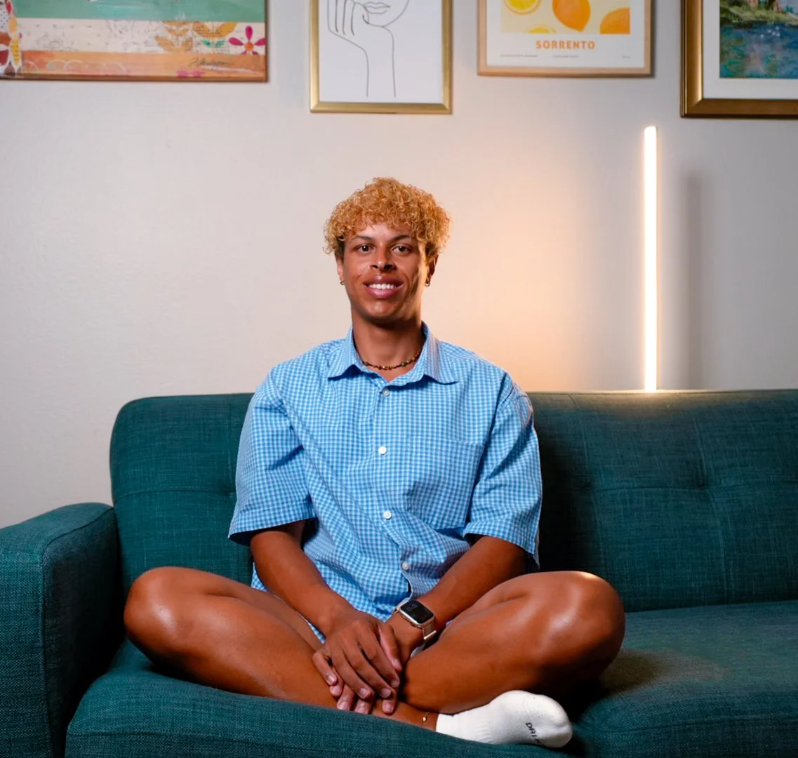 A person sitting cross-legged on a couch, smiling, wearing a blue checkered shirt, white socks, with artwork hanging on the wall behind them.