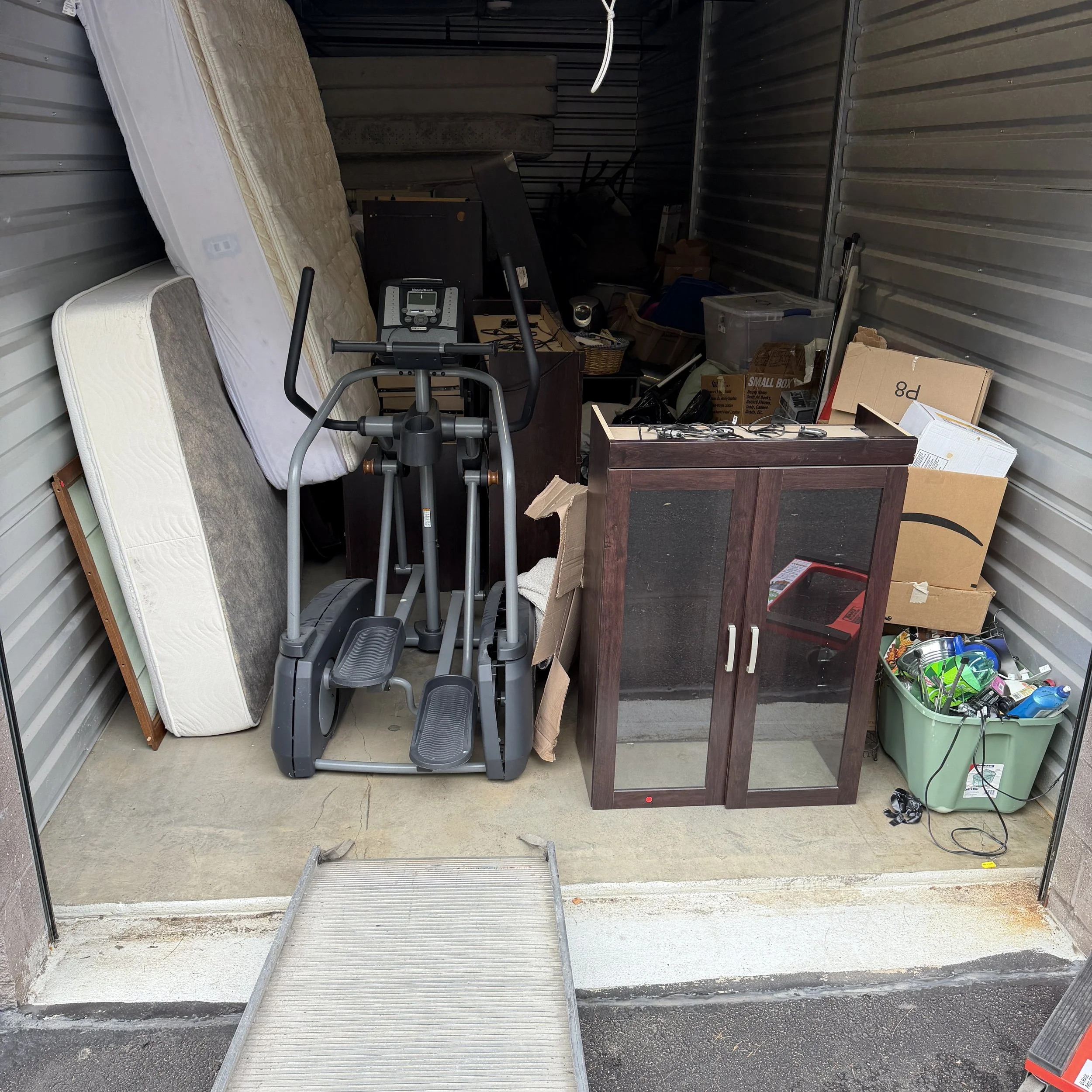 Inside a storage unit with miscellaneous items including a used mattress, a treadmill, a wooden desk, boxes, and electronic cords.