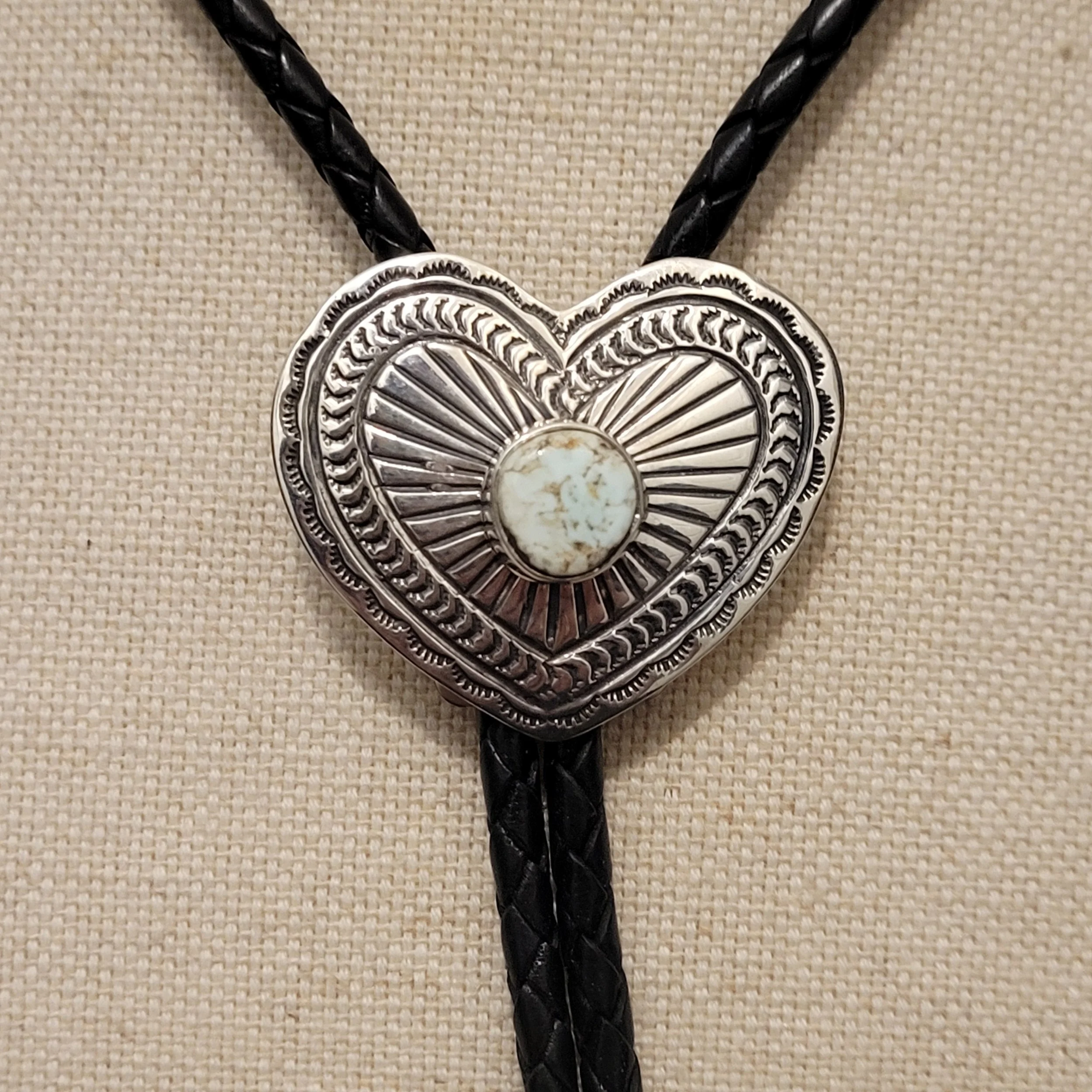 A silver and black decorative heart-shaped pendant with a white stone in the center, hanging from a black twisted cord, on a beige textured background.