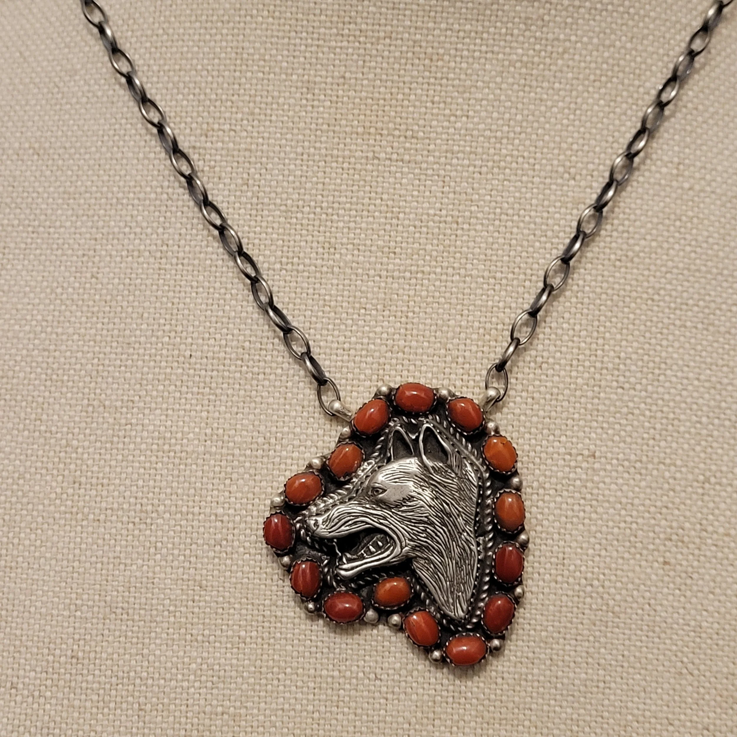 A necklace with a large silver pendant featuring a detailed design of a woman's face, surrounded by orange beads, silver beads, and a silver chain on a beige textured background.