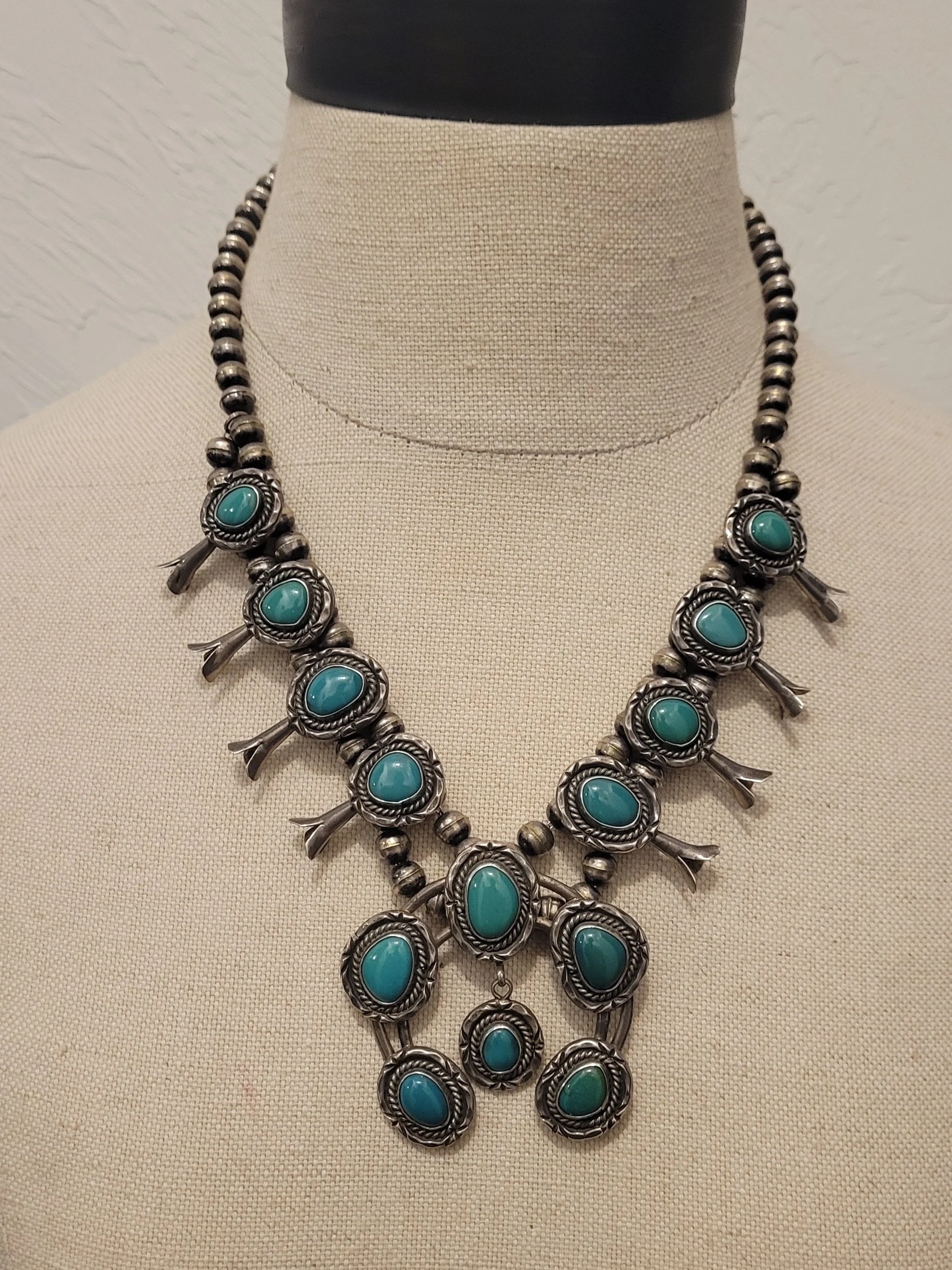 A vintage-style silver necklace with turquoise stones displayed on a beige fabric bust.