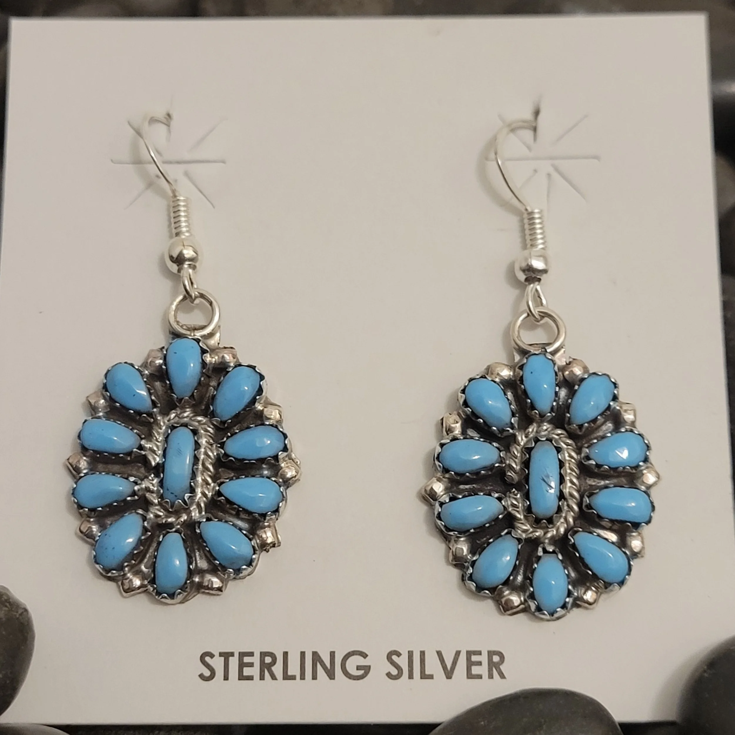 A pair of silver earrings with light blue stones, displayed on a white card labeled 'Sterling Silver'. The earrings are hanging with attached hooks and decorative elements surrounding the stones.