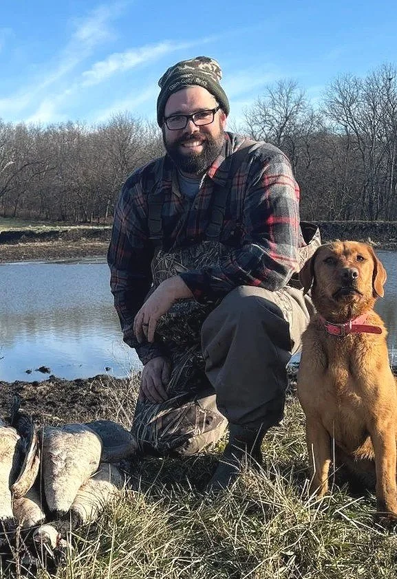 A man with glasses, a beard, and wearing outdoor camo clothing and a plaid shirt is kneeling next to a brown hunting dog with a red collar, near a body of water and trees in the background.