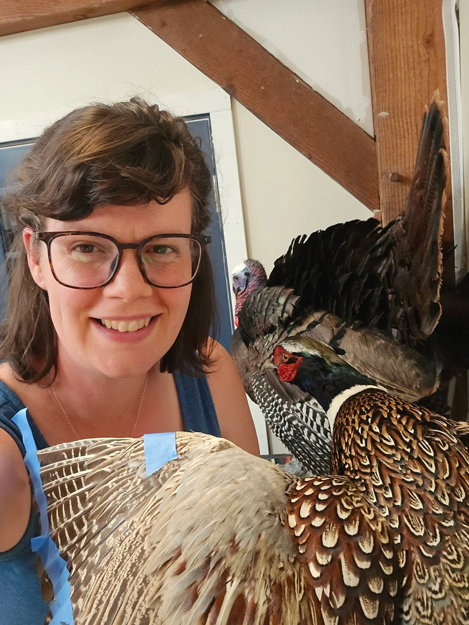 A woman with glasses smiling next to a display of various colorful birds, including a turkey with brown, black, and white feathers, and a colorful pheasant with red, green, and black plumage.