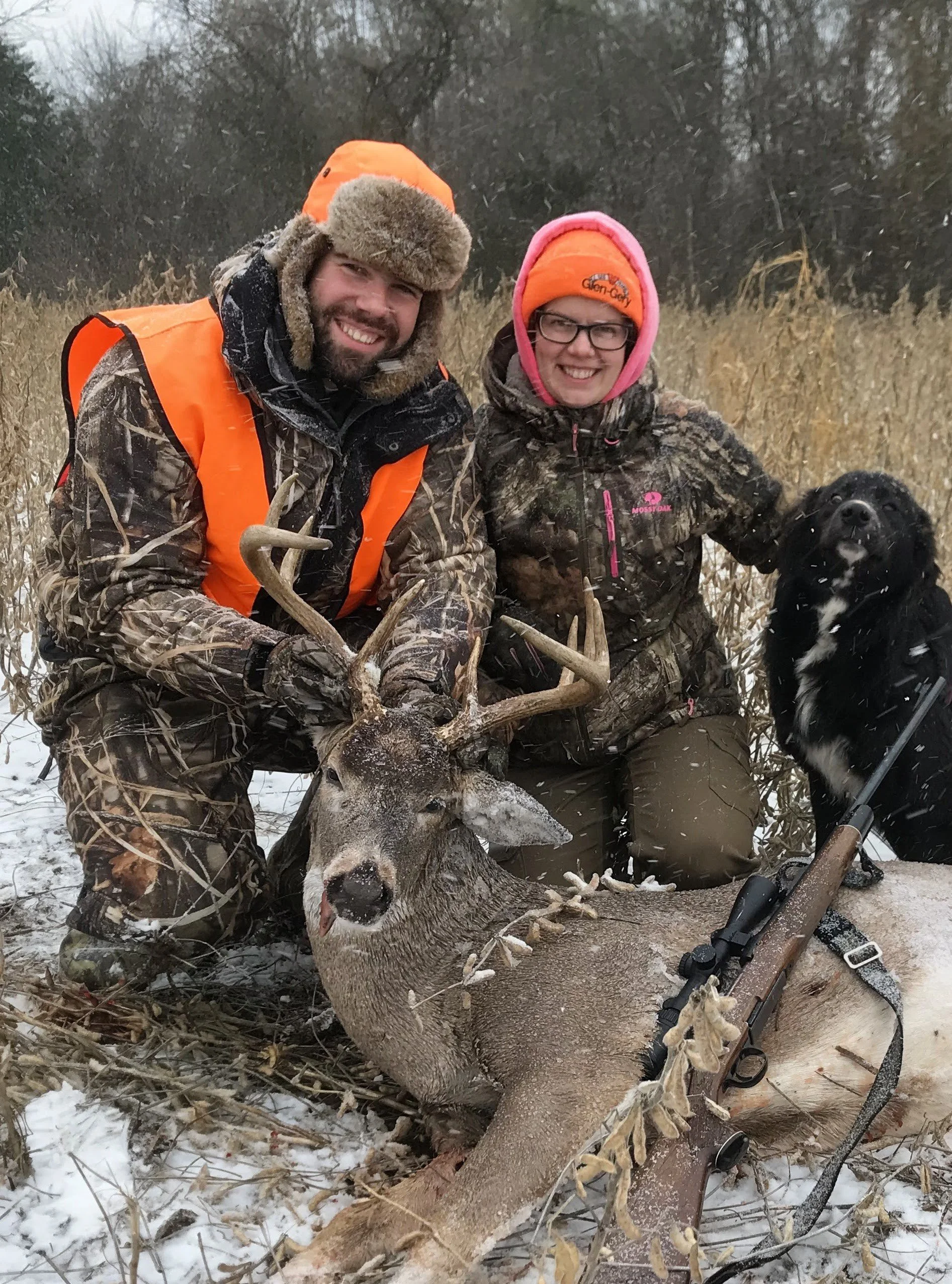 Two hunters in camouflage clothing and orange hats pose with a deer they hunted in a snowy field with tall grass and trees in the background. The man on the left is holding the deer's antlers, and the woman on the right is smiling. There is also a rifle resting on the ground near the deer.