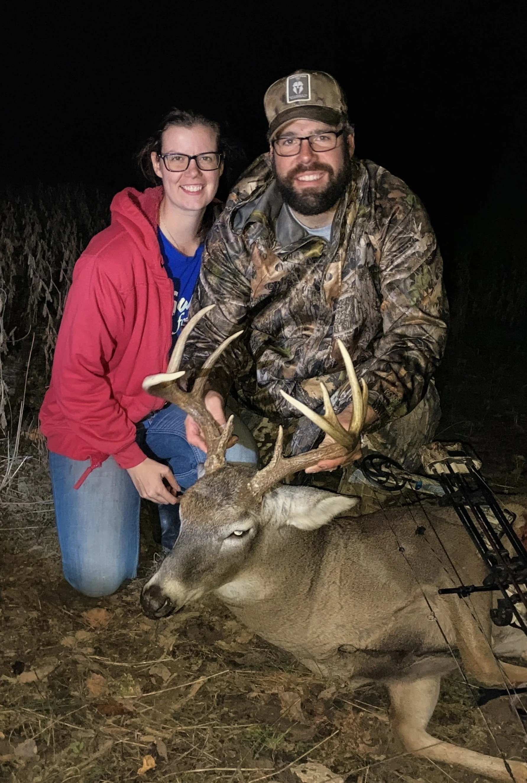 A woman and a man with glasses and beards, smiling and holding the antlers of a large buck in a wooded outdoor location at night. The woman is wearing a red jacket and glasses, and the man is dressed in camouflage, also with a hat and glasses. There's a bow and arrow lying on the ground next to them.