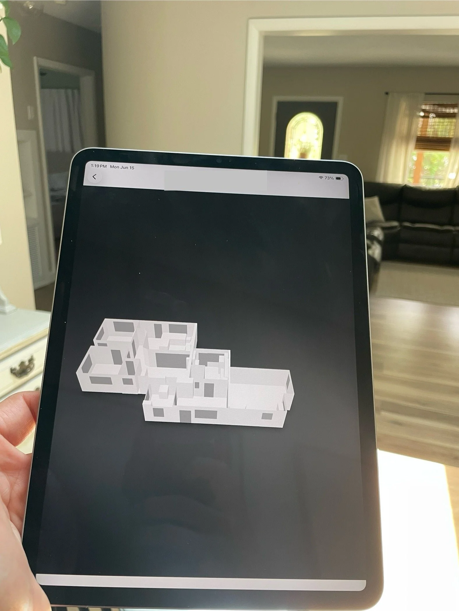 A person holding a tablet with a 3D building floor plan displayed on the screen, in a living room with a dark couch, windows with curtains, and an entry door in the background.