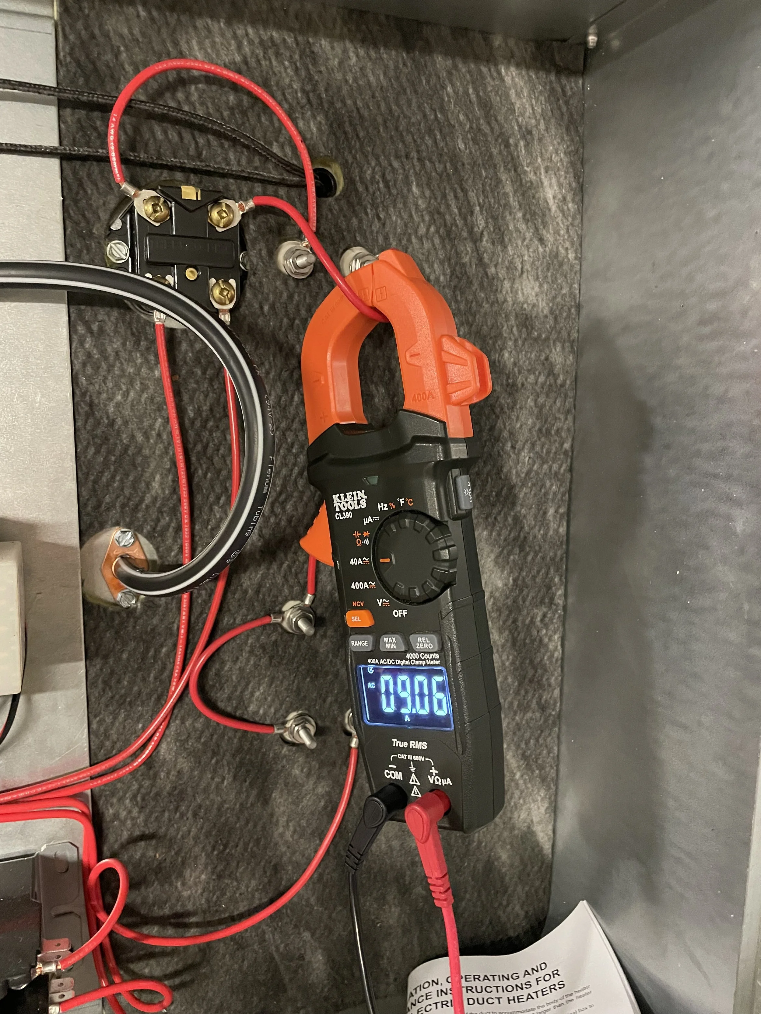 Digital clamp meter measuring electrical current, clipped onto wires connected to electronic components, with manila paper partially visible at bottom.