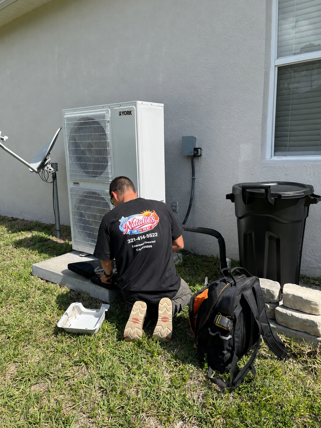 Technician working on an outdoor air conditioning unit beside a house, with tools and a backpack nearby.
