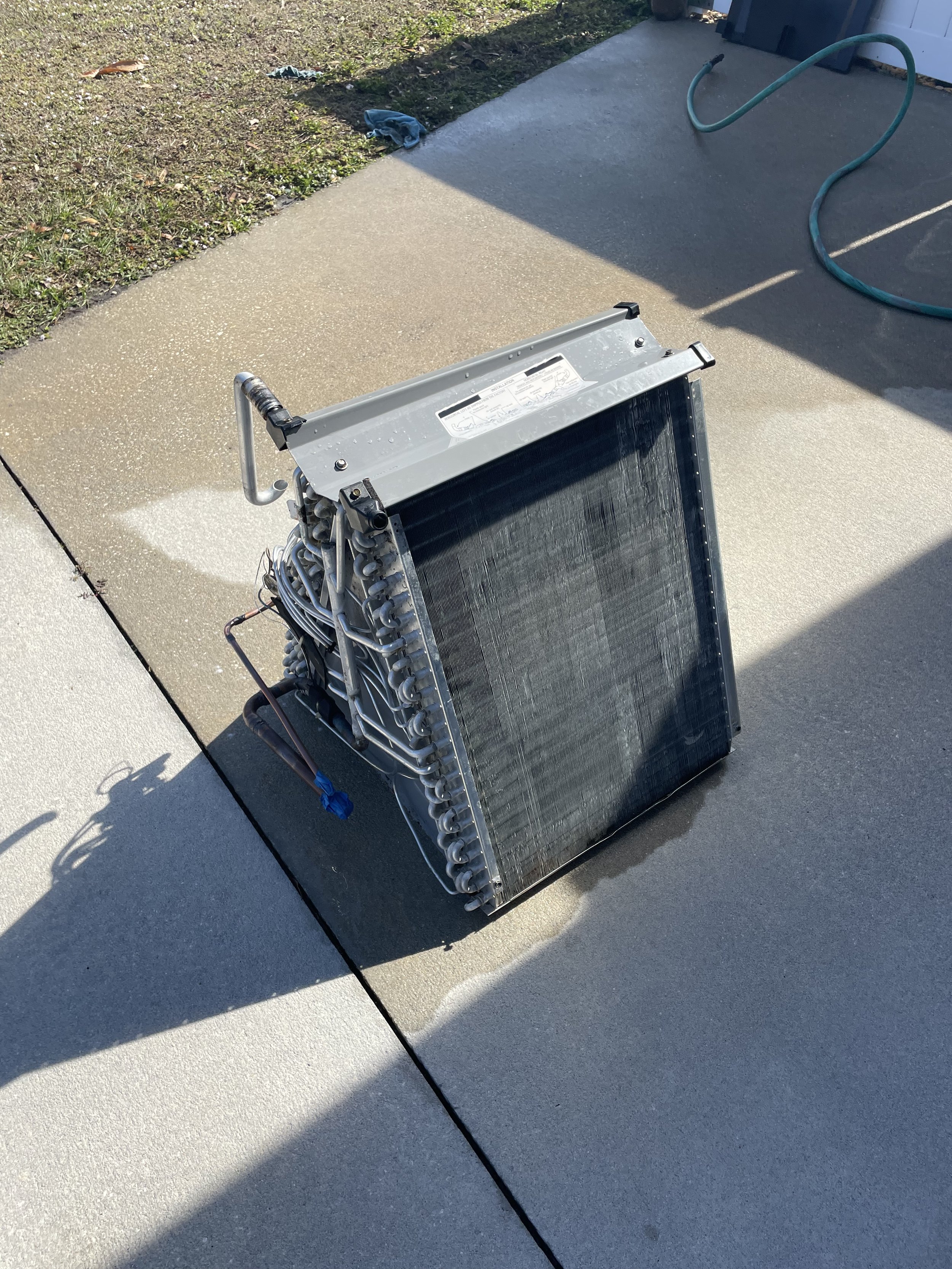 An air conditioning evaporator with metal frame and tubing lies on a concrete driveway. A garden hose is used to clean the coil. Lineset connections are taped shut so water and moister don't enter the evaporator coil