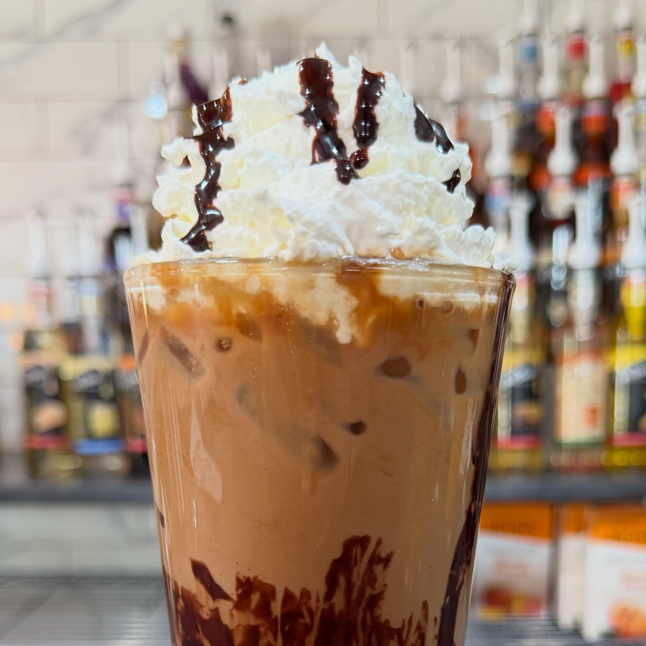 A close-up of a glass of iced coffee topped with whipped cream and chocolate syrup, with a blurred background of shelves with various bottles.