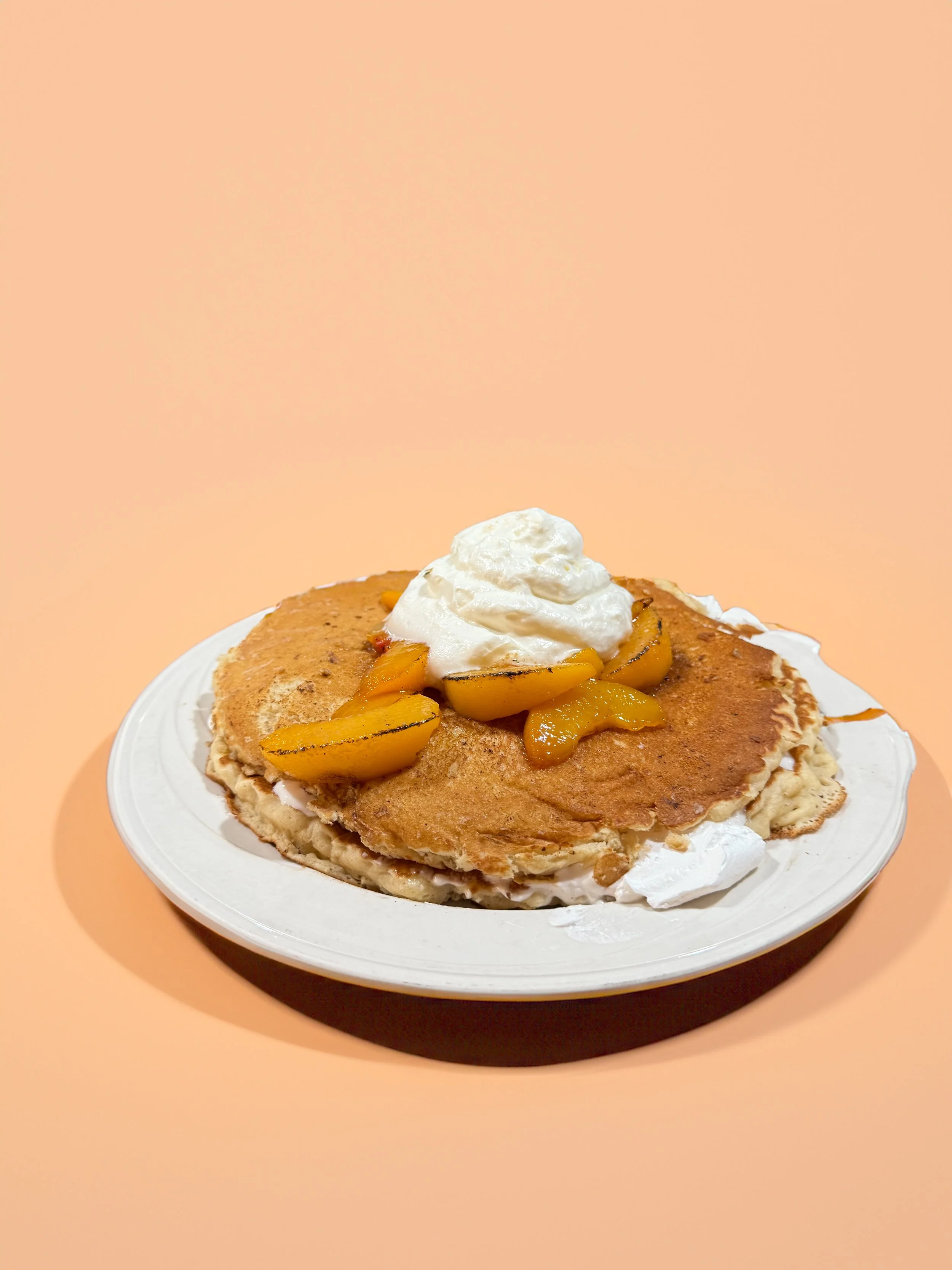 Close-up of a plate with a stack of pancakes topped with whipped cream and peach slices, on a peach-colored background.