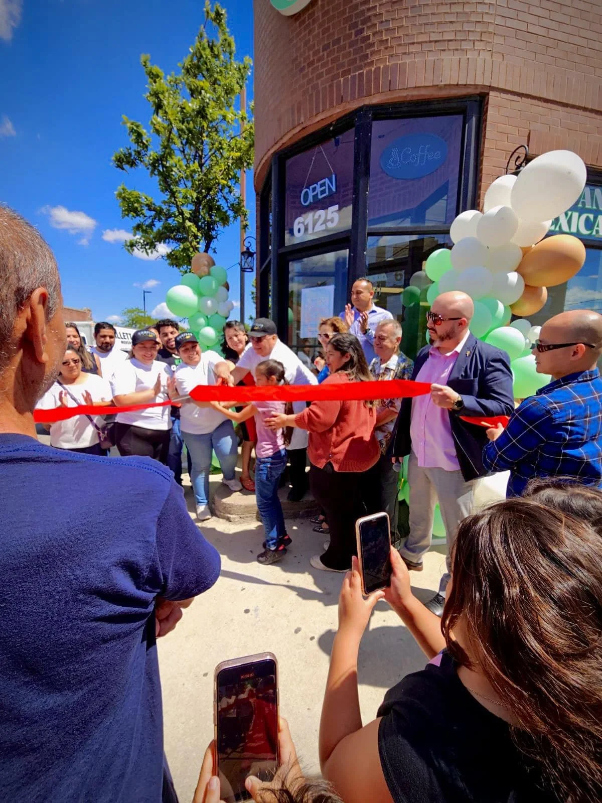 People gathered outside a brick building with balloons in green, white, and gold for a ribbon-cutting ceremony. A woman is cutting a red ribbon while others, including children and officials, cheer and take photos. The building has the address 6125 and an 'Open' sign in the window.