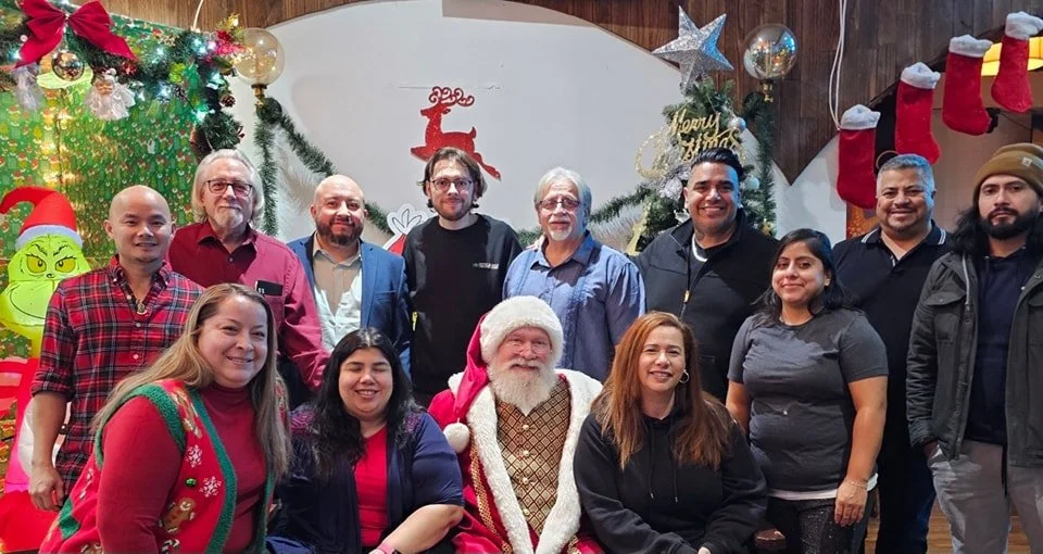 Group of people gathered around Santa Claus for a Christmas celebration with festive decorations including stockings, Christmas trees, ornaments, and a reindeer cutout.