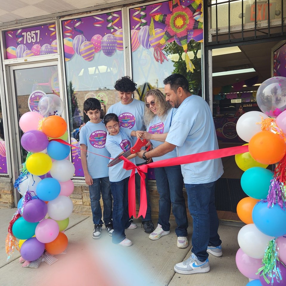 A group of five people, including children and adults, are participating in a ribbon cutting ceremony outside a decorated storefront for a new business or event. They are surrounded by colorful balloons and festive decorations, with a person using scissors to cut a red ribbon.
