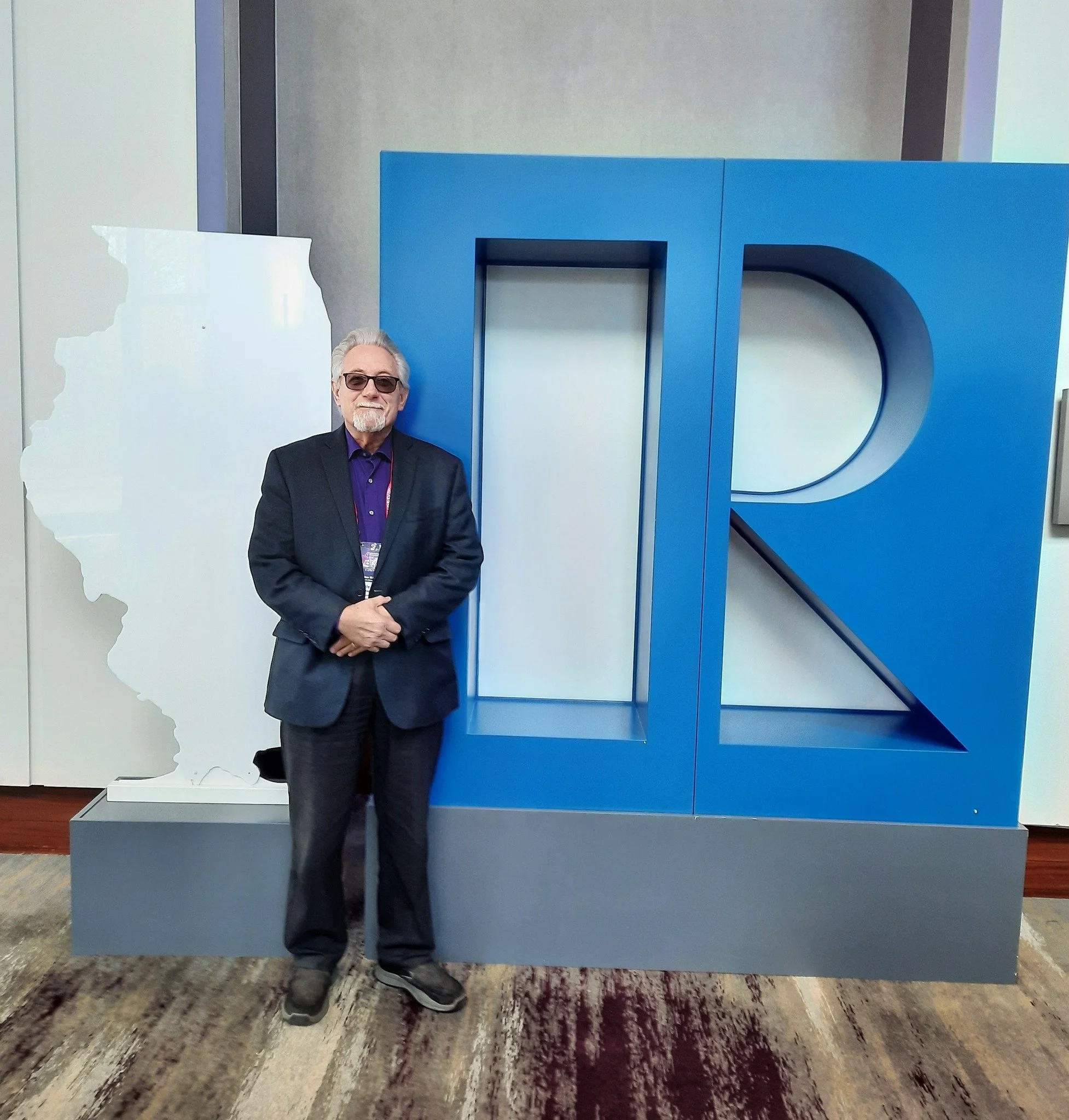 A man with gray hair and beard, wearing glasses, a dark suit, purple shirt, and standing with his hands clasped in front of him, is positioned in front of a large blue structure with the letters 'D' and 'E' visible.