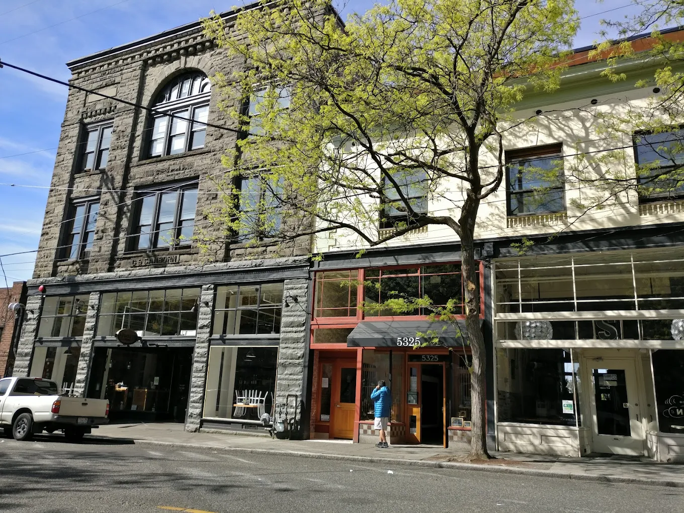 Street view of a historic multi-story building with large windows, a gray stone facade, and a business at the ground level, with a person standing outside. A tree with budding green leaves partially obscures the building.