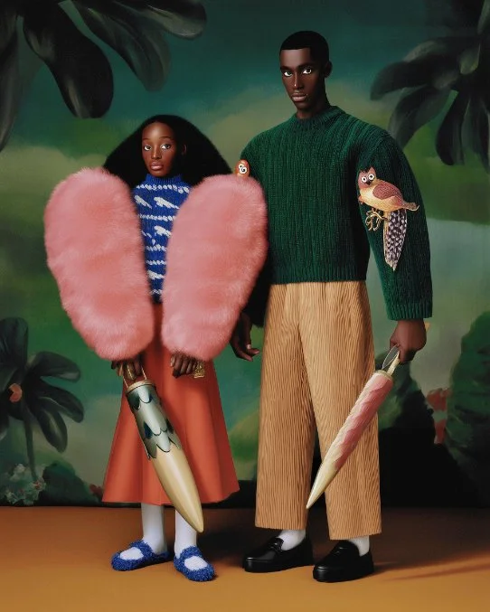 Two models standing in front of a jungle-themed backdrop, wearing colorful and eclectic clothing. The female model is dressed in a blue and white patterned top, a brown skirt, bright pink fluffy jacket, and fuzzy slippers. She holds a large umbrella with a cartoon face design. The male model is wearing a green textured sweater with animal patches, striped pants, and black shoes, and holds a large umbrella with a pink and beige pattern.