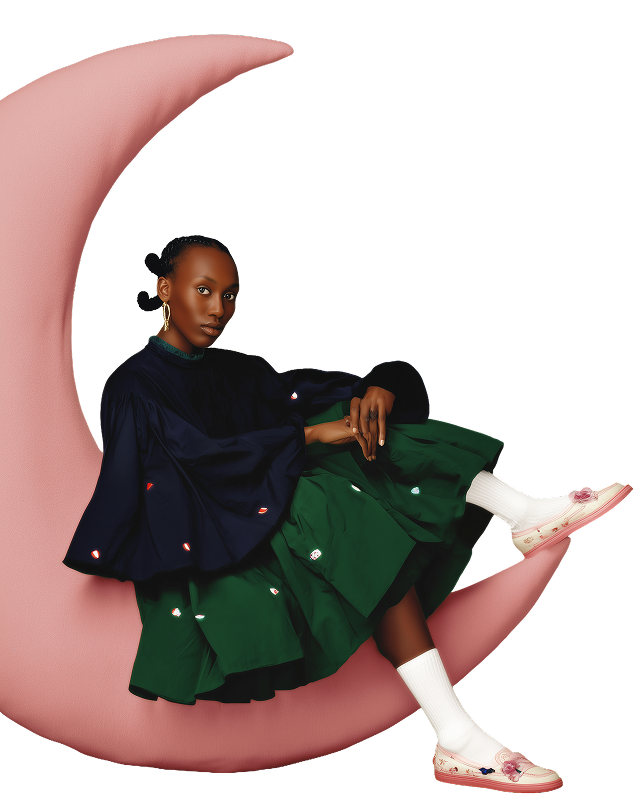 A woman sitting on a pink crescent moon prop, wearing a black top with red and white embellishments, green pleated skirt, white socks, and pink shoes with floral decorations, against a black background.