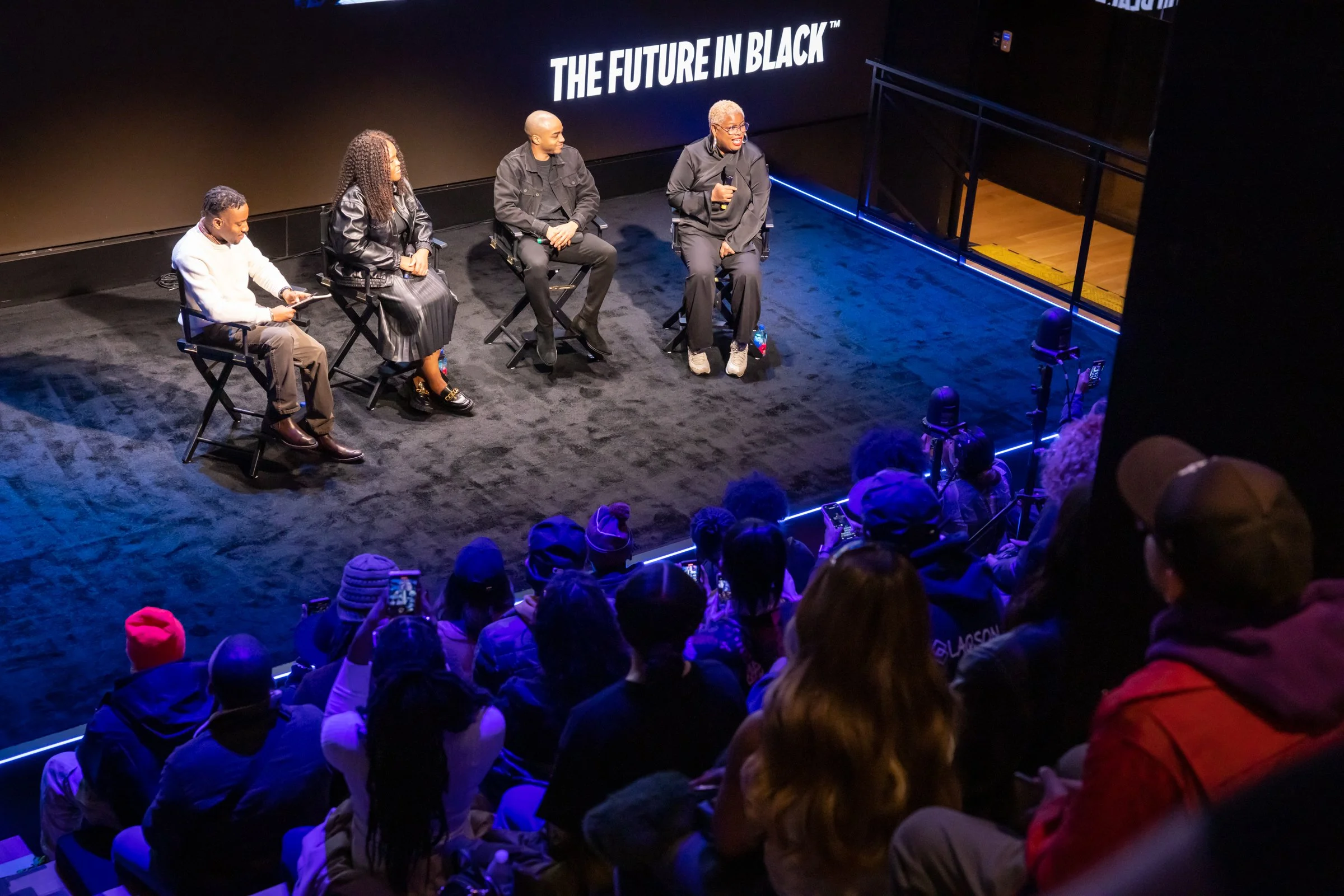 A panel of four people sitting on chairs on a stage, with a large screen behind them displaying 'THE FUTURE IN BLACK™'; an audience watching and taking photos.